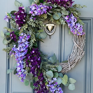 May include: A purple and blue lilac wreath with eucalyptus leaves on a gray door. The wreath is made of grapevine and has a silver door knocker with the text "J & T Pagliaro" on it.