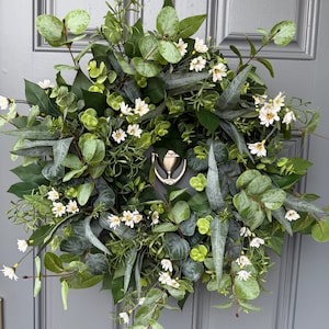 May include: A lush, green wreath with a variety of foliage and small white flowers. The wreath is adorned with eucalyptus leaves, creating a natural and inviting aesthetic. A silver door knocker is visible in the center.