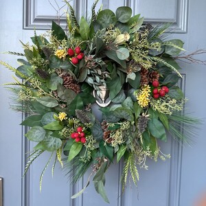 May include: A lush, round wreath featuring a variety of green foliage, including eucalyptus and pine. Accents of red berries, yellow flowers, and pinecones add color and texture. A decorative element is visible in the center.
