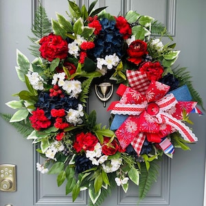 May include: A patriotic wreath with red, white, and blue flowers and greenery. The wreath features red roses, white blossoms, and blue hydrangeas, accented by a large red bow with a button center. The wreath is displayed on a gray door.
