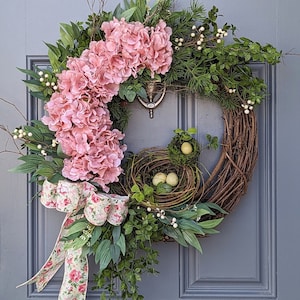 May include: A pink and green spring wreath with a bird's nest and eggs. The wreath is made of grapevine and has a floral ribbon.