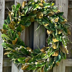 May include: A circular wreath composed of green magnolia leaves, accented with brown leaves and small white flowers. The wreath is displayed against a weathered wooden fence, creating a natural and rustic aesthetic.