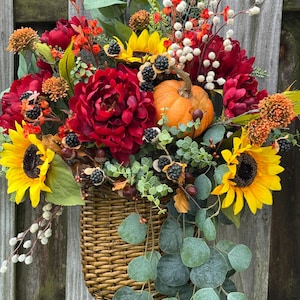 May include: A woven basket overflowing with a vibrant autumn floral arrangement. The arrangement includes sunflowers, red peonies, an orange pumpkin, and berries. Green eucalyptus leaves cascade down the side, creating a warm, seasonal display.