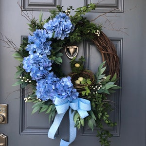 May include: A handcrafted wreath featuring blue hydrangea flowers, greenery, and a bird's nest with eggs. A light blue polka dot bow adds a decorative touch. The wreath is displayed on a gray door.