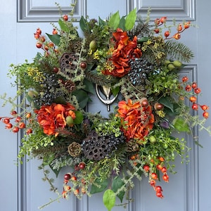 May include: A circular wreath featuring deep orange hydrangea blossoms, dark pine needles, green foliage, brown pinecones, dried lotus seed pods, and clusters of small red berries. The arrangement is mounted on a gray door with a silver door knocker.