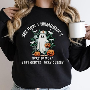 May include: Black sweatshirt with a white ghost wearing a nurse's hat and holding a pumpkin. The text on the sweatshirt reads "See how I immunize? Very demure, very gentle, very cutesy."