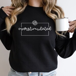 May include: Black sweatshirt with white text that reads "overstimulated" inside a white rectangle. A white doodle of a tangled line is above the text.
