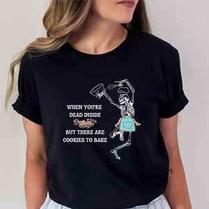 May include: Black t-shirt featuring a skeleton chef with a mixing bowl and whisk, wearing an apron. The shirt includes the text: "When you're dead inside but there are cookies to bake."