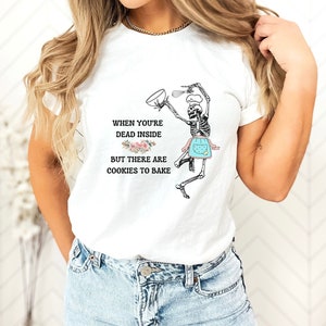 May include: White t-shirt with a humorous graphic of a skeleton baking cookies. The skeleton is wearing a chef's hat and apron, holding a bowl and whisk. Text reads: "When you're dead inside but there are cookies to bake."