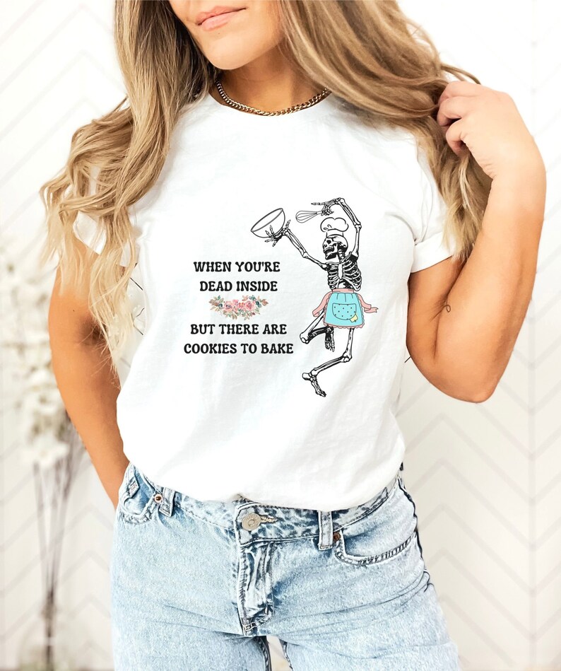 May include: White t-shirt with a humorous graphic of a skeleton baking cookies. The skeleton is wearing a chef's hat and apron, holding a bowl and whisk. Text reads: "When you're dead inside but there are cookies to bake."