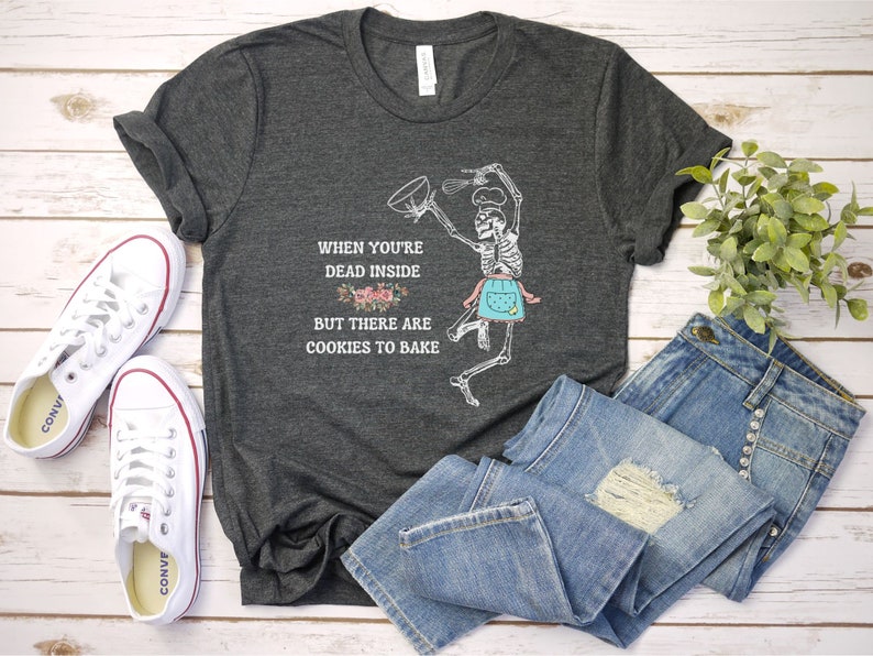May include: Dark gray t-shirt with a skeleton chef graphic, wearing a blue apron and chef's hat, holding a whisk and bowl. Text reads: "When you're dead inside but there are cookies to bake." White sneakers and blue jeans are also in the image.