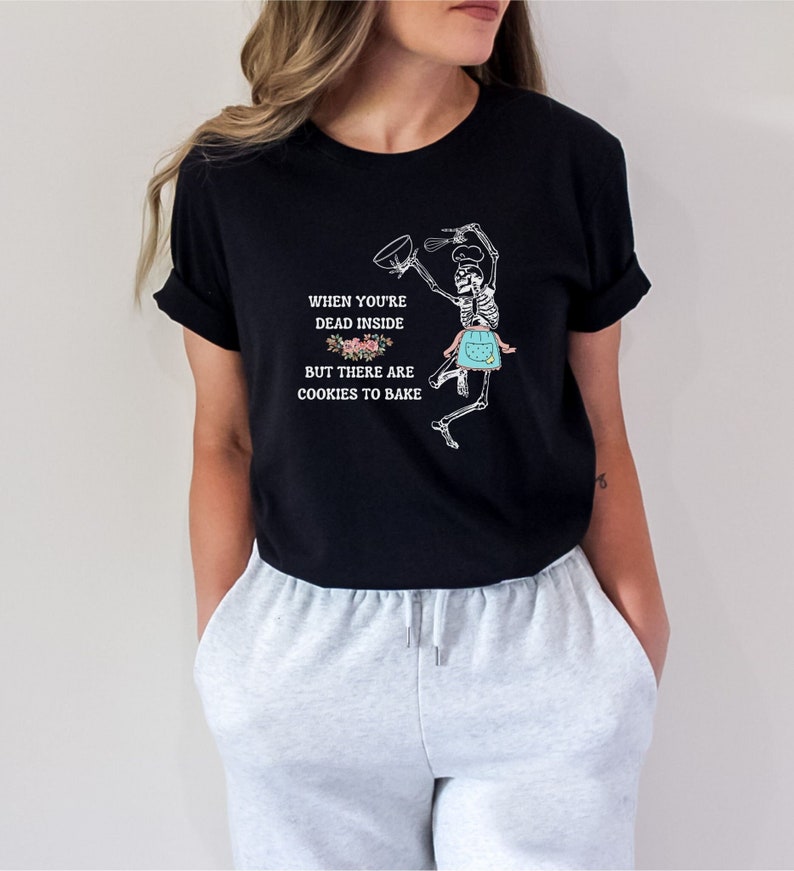 May include: Black t-shirt featuring a skeleton chef with a mixing bowl and whisk, wearing an apron. The shirt includes the text: "When you're dead inside but there are cookies to bake."