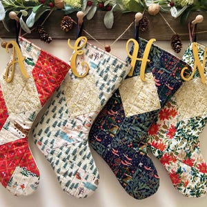 May include: Four quilted Christmas stockings with gold wooden letter ornaments. The stockings feature various patterns, including red and white geometric designs, forest scenes, and floral prints. Each stocking is hung from a wooden rod.