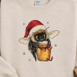 May include: A sand-colored crewneck sweatshirt featuring a graphic of a fuzzy bumblebee wearing a red Santa hat. The bee is holding a glass jar of golden honey tied with a red ribbon, surrounded by small black snowflake illustrations.