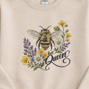 May include: A beige crewneck sweatshirt featuring a central graphic of a bumblebee adorned with small yellow and white flowers. The bee is surrounded by lavender sprigs, yellow buttercups, and white daisies, with the word Queen in black script.