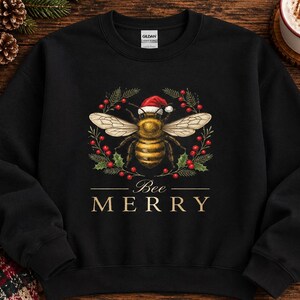 May include: A black crewneck sweatshirt featuring a graphic of a bee wearing a red and white Santa hat, surrounded by a wreath of green pine needles and red berries. Below the bee, the text reads Bee MERRY in gold, capitalized lettering.