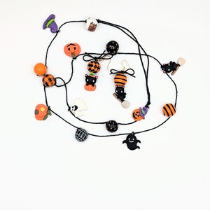May include: A black necklace with orange and black Halloween themed charms. The charms include pumpkins, ghosts, bats, and cats. The necklace is strung on a black cord and has a lobster clasp closure.