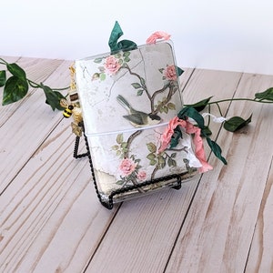May include: A white and green floral journal with a bird and a bee charm. The journal is on a black metal stand and has a clear plastic cover.