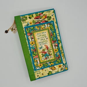May include: A rectangular journal with a green polka dot spine and a cover featuring illustrations of mushrooms, butterflies, and clover. The center panel displays the text, May your troubles be few and your blessings be many, with a gold metal charm.