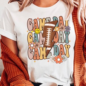 以下が含まれることがあります： カラフルな文字で「ゲームデー」と書かれたレトロなデザインの白いTシャツ。フットボールと花が描かれています。
