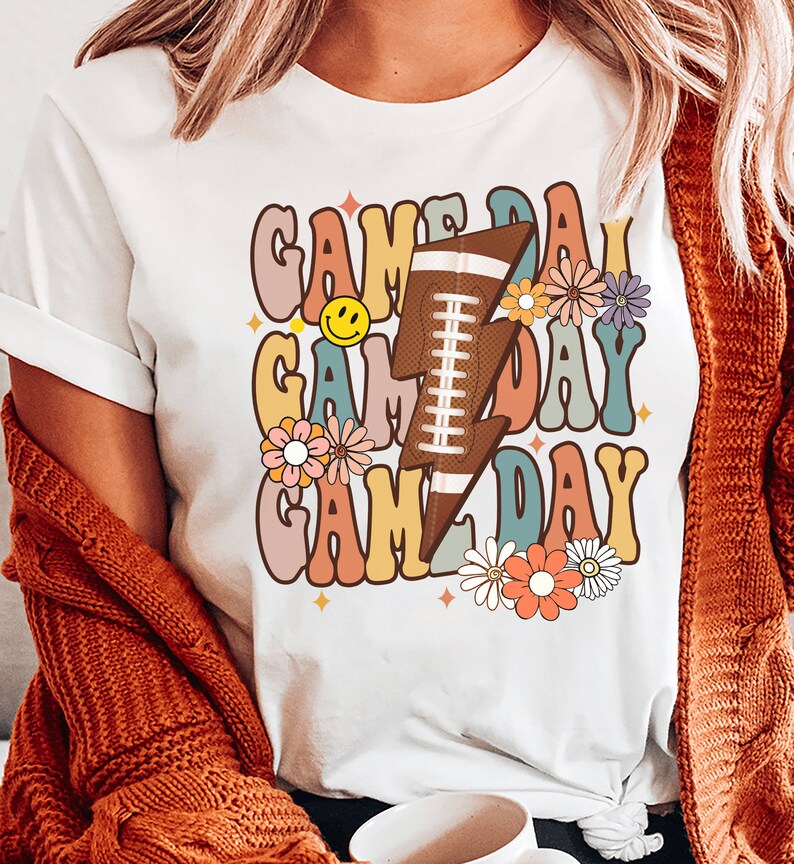 以下が含まれることがあります： カラフルな文字で「ゲームデー」と書かれたレトロなデザインの白いTシャツ。フットボールと花が描かれています。