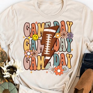 以下が含まれることがあります： レトロなフォントで「Game Day」という文字が3回繰り返されたベージュのTシャツ。文字の周りは花と、フットボールが入った稲妻で囲まれています。