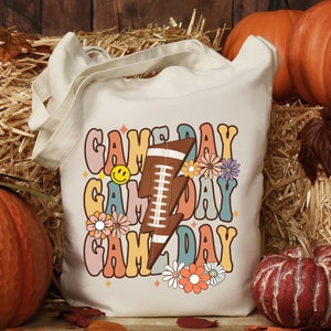 以下が含まれることがあります： レトロなデザインが施された白いキャンバス地のトートバッグ。"GAME DAY"という文字が3回、異なる色とフォントで繰り返されています。デザインの中央には、フットボールが入ったスタイリッシュな稲妻が描かれています。デザインはカラフルな花で囲まれています。