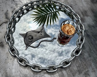 May include: A silver chain tray with a mirrored surface reflecting a palm leaf, a glass of iced coffee, and a silver necklace.
