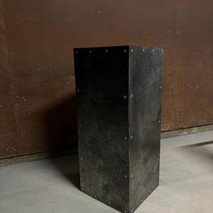 May include: A tall, rectangular metal pedestal with a dark gray finish. The pedestal has a slightly textured surface and is secured with small rivets.