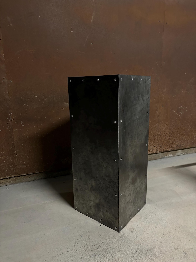 May include: A tall, rectangular metal pedestal with a dark gray finish. The pedestal has a slightly textured surface and is secured with small rivets.