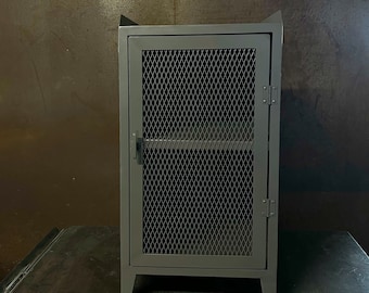 May include: A gray metal cabinet with a wire mesh door and a shelf inside. The cabinet has a simple design with a rectangular shape and four legs.