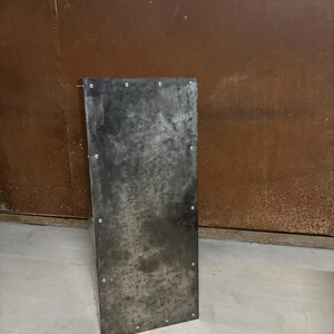 May include: A tall, rectangular metal stand with a dark gray finish and a slightly textured surface. The stand has a series of small, evenly spaced rivets around the edges.