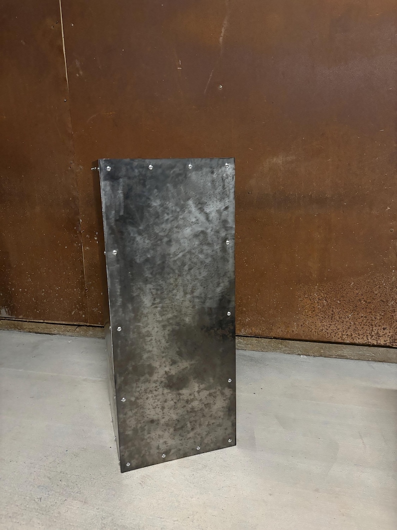 May include: A tall, rectangular metal stand with a dark gray finish and a slightly textured surface. The stand has a series of small, evenly spaced rivets around the edges.
