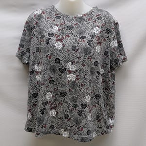 May include: Gray short-sleeved shirt with a floral pattern. The shirt features a round neckline and a repeating design of white, black, and burgundy flowers and leaves. The background has a subtle, textured pattern.