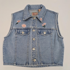 May include: Light blue denim vest with embroidered pink flowers. The vest has a collared neckline, two front pockets, and silver-tone button closures. The brand name "Kikomo" is visible on the vest.