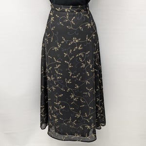 May include: A black midi skirt with a flowing silhouette. The skirt features a delicate pattern of beige and blue leaves and vines. The fabric appears to be lightweight and semi-sheer, suitable for warmer weather.