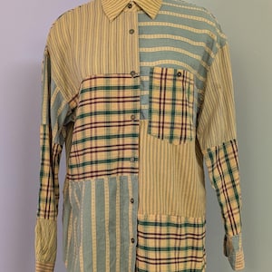 May include: A long-sleeve button-down shirt featuring a patchwork design of mustard yellow, sage green, and dark maroon fabrics. The shirt displays alternating panels of vertical stripes and plaid patterns, including a single chest pocket on the left.