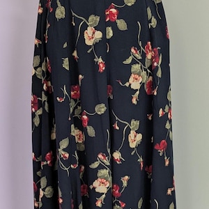 May include: A floor-length skirt in a dark navy blue color featuring a repeating floral pattern of red and cream-colored blossoms with muted green leaves and thin stems. The skirt has a fitted waistband and a flowing, pleated silhouette.
