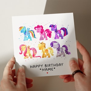 May include: A white greeting card with watercolor illustrations of colorful ponies. The card reads "HAPPY BIRTHDAY *NAME*" in black text, with a small red heart. The ponies are in shades of blue, pink, purple, yellow, and orange.