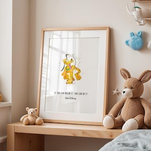 Op de afbeelding: Een ingelijste print met een aquarel illustratie van Pluto in gele en oranje tinten. De print bevat de quote "IF YOU CAN DREAM IT, YOU CAN DO IT." van Walt Disney. Het kunstwerk wordt tentoongesteld op een houten plank met knuffels.