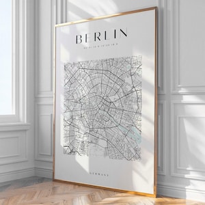 Op de afbeelding: Een ingelijste print van een kaart van Berlijn, Duitsland, met de naam van de stad en de coördinaten bovenaan. De kaart is een gedetailleerde zwart-wit illustratie van de straten en waterwegen van de stad. De lijst is goudkleurig.