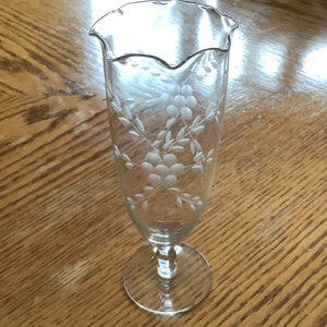 May include: A clear glass vase with a scalloped rim and etched floral design. The vase has a tall, slender body and a round base.