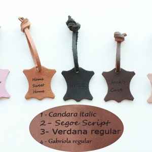 Könnte beinhalten: Fünf Leder-Schlüsselanhänger in verschiedenen Farben und mit unterschiedlichem Text. Der Text auf den Schlüsselanhängern lautet: "Alindas Zimmer", "Home Sweet Home", "NEDE", "Jacks Höhle", und "Saralys Küche". Die Schlüsselanhänger sind mit verschiedenen Schriftarten beschriftet: Candara kursiv, Segoe Script, Verdana regular, und Gabriola regular.