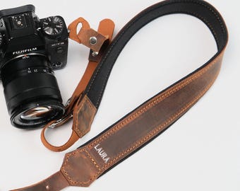 Może przedstawiać: Czarny aparat Fujifilm z obiektywem i brązowym skórzanym paskiem do aparatu. Pasek ma czarny spód, a na nim nadrukowane jest imię "LAURA". Pasek jest połączony z aparatem za pomocą metalowego osprzętu.