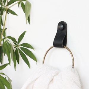 Puede incluir: Anillo para toalla de cuero negro con un anillo de latón. El anillo está montado en una pared blanca con un tornillo negro.