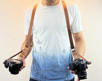 May include: A person wearing a white t-shirt with a blue and white pattern, holding two black cameras with brown leather straps. The straps are connected to a brown leather camera strap that is worn around the neck.