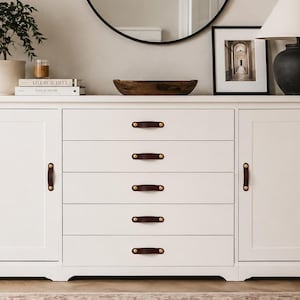 Könnte beinhalten: Ein weißes Sideboard aus Holz mit zwei Seitenschränken und fünf mittigen Schubladen. Jede Schublade und Schranktür ist mit einem braunen Ledergriff ausgestattet, der durch messingfarbene Beschläge befestigt ist. Das Möbel hat eine flache Oberfläche.