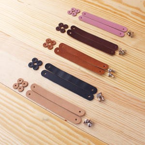 May include: An overhead shot displays sets of leather straps in various colors: pink, brown, tan, and navy blue. Each set includes two straps, rivets, and matching washers. The items are arranged on a light-colored wooden surface.