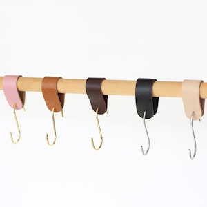 Puede incluir: Cinco ganchos envueltos en cuero cuelgan de una barra de madera. Los ganchos son de color rosa, marrón, negro y beige. Los ganchos son de metal y tienen un acabado dorado.