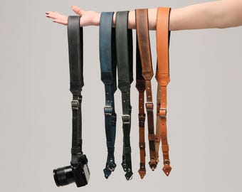 Puede incluir: Una colección de correas de cámara de cuero en varios colores, incluyendo negro, verde azulado, marrón y bronceado. Cada correa se muestra, con una cámara adjunta a una de las correas negras. Las correas tienen hebillas ajustables.